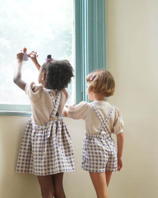 Two children wearing soor ploom's enid shortall in blueberry gingham print, made from soft cotton, gaze out a window. One holds up a doll.