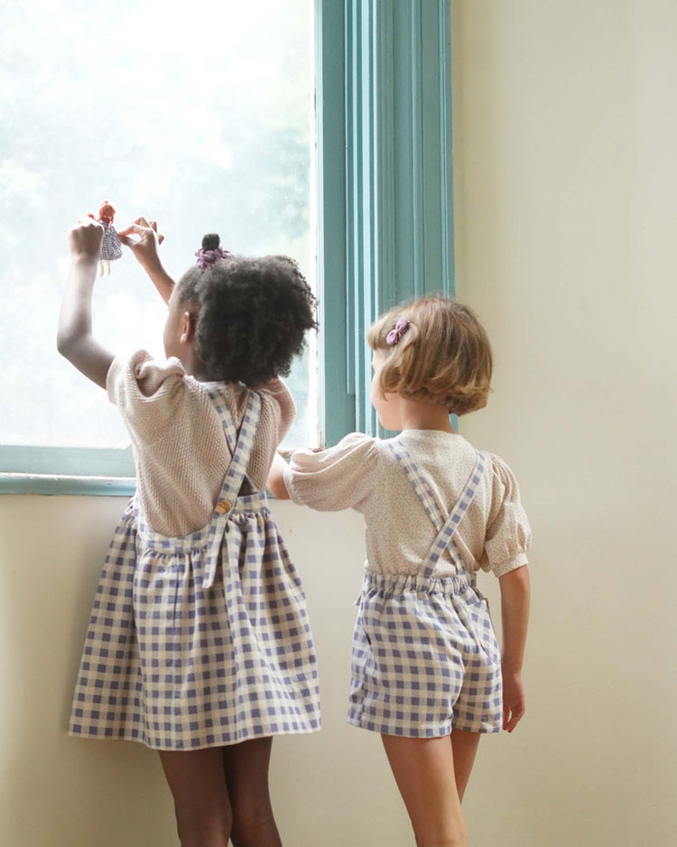 Two children wearing soor ploom's enid shortall in blueberry gingham print, made from soft cotton, gaze out a window. One holds up a doll.