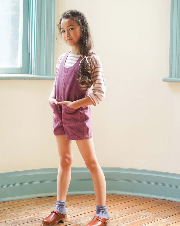 A young girl stands indoors on a wooden floor near a turquoise-trimmed window, wearing a striped cotton shirt and the "delphine shortall in zinnia" by Soor Ploom with brown clogs. Her long braided hair cascades down as a vase of zinnias enhances the vibrant scene.