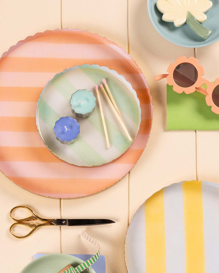 The meri meri pastel striped large accessory tray is arranged on a cream-colored surface, alongside pastel candles, long matches, flower-shaped sunglasses, scissors, and a green napkin.