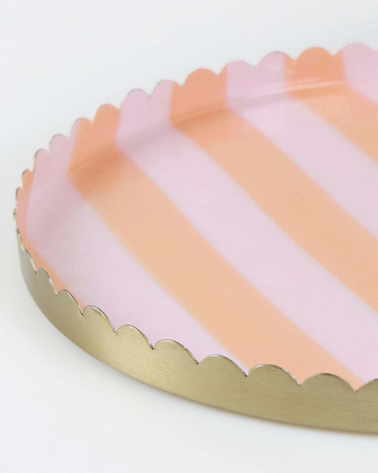 The meri meri pastel striped large accessory tray is a round metal tray with scalloped edges, an enamel finish, and alternating pink and peach stripes.