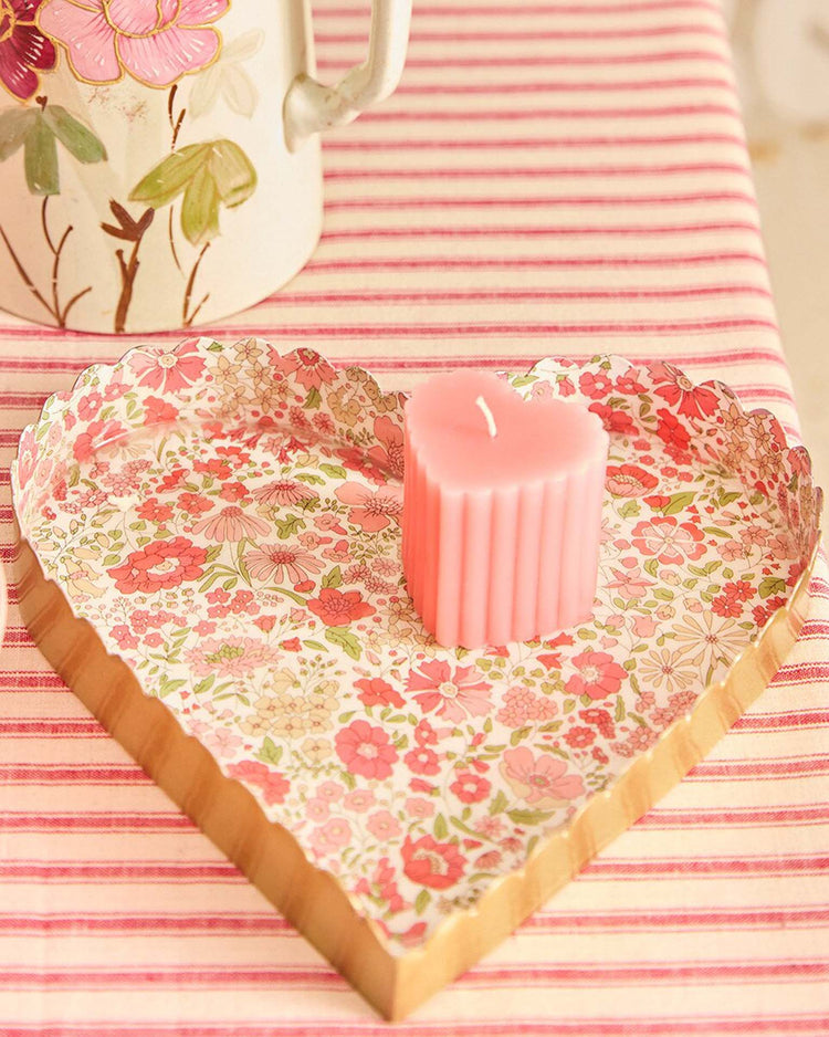 A pink heart-shaped candle rests on the meri meri x liberty heart large accessory tray by meri meri, featuring a Liberty floral print and gold edge—an ideal Valentine’s gift set atop a pink and white striped tablecloth.