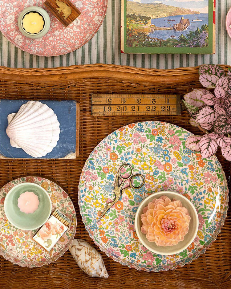 The meri meri x liberty round medium accessory tray by meri meri displays floral plates, a ruler, seashells, vintage books, scissors, a pink flower in a bowl, two candles, and a small pink plant on a striped cloth background.