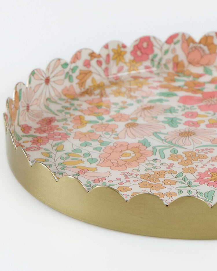 The meri meri x liberty round small accessory tray by meri meri features a colorful Liberty floral pattern with pink, orange, yellow, and green flowers and leaves on enamel metal with scalloped gold edges.