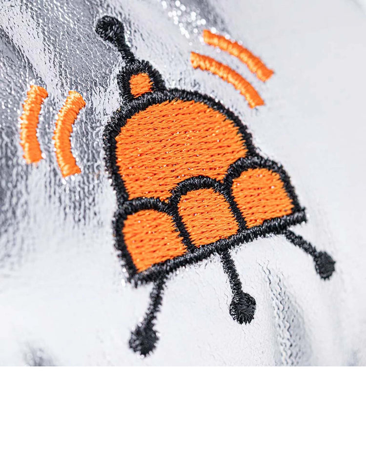 Close-up of an embroidered orange and black dome with curved signal-like lines on shiny silver fabric—capturing the playful charm of the amuseables jellysaucer by Jellycat.