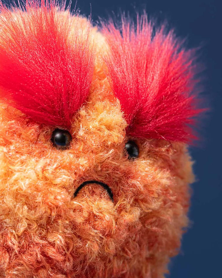 A close-up of a fuzzy orange Jellycat amuseables planet mars plush toy with bright red bushy eyebrows and a sad expression, set against a dark blue background.