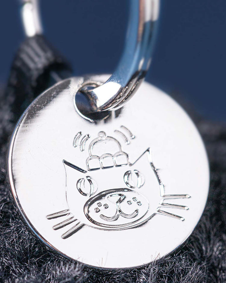 A close-up of the Jellycat amuseables black hole bag charm: a shiny, round metal tag on a keyring, engraved with a cartoon cat face in a beanie—an ideal unique physics gift or quirky bag accessory—shown against a dark textured background.