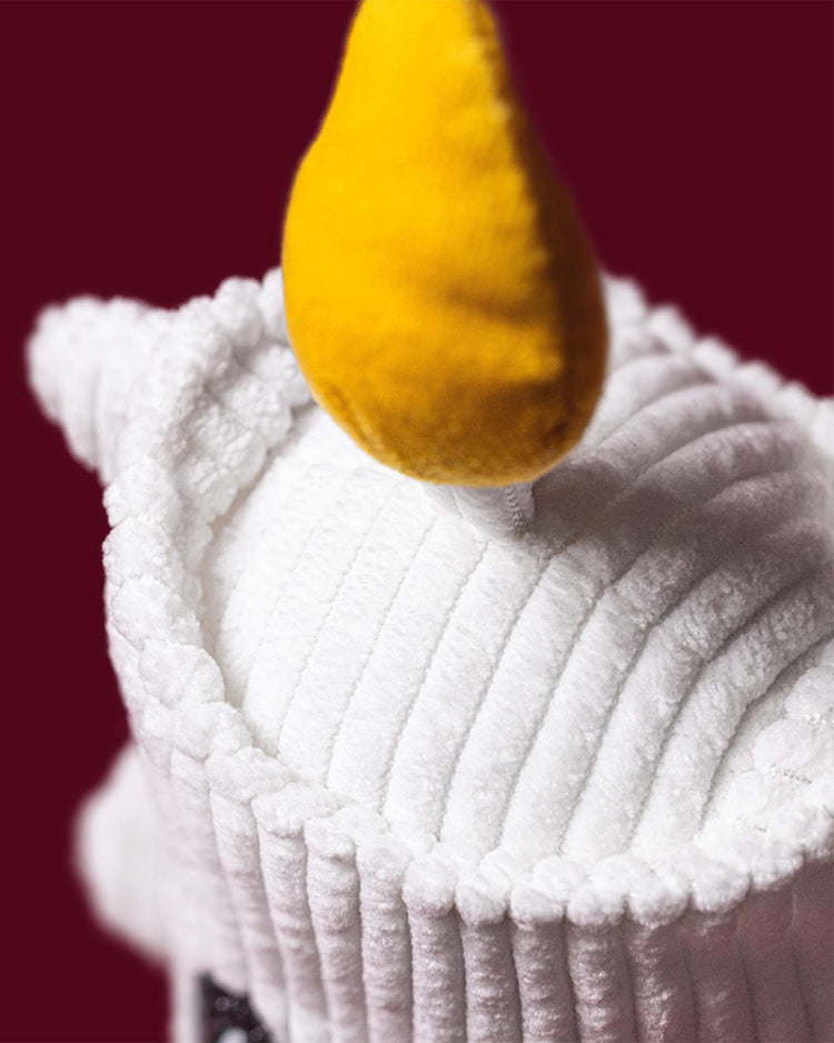 Close-up of the Jellycat Amuseables Flickerty Candle plush, featuring a white cupcake-inspired design with a soft, yellow flame on top, set against a solid burgundy background—a charming pick for any Halloween party planner curating spooktacular tablescapes.
