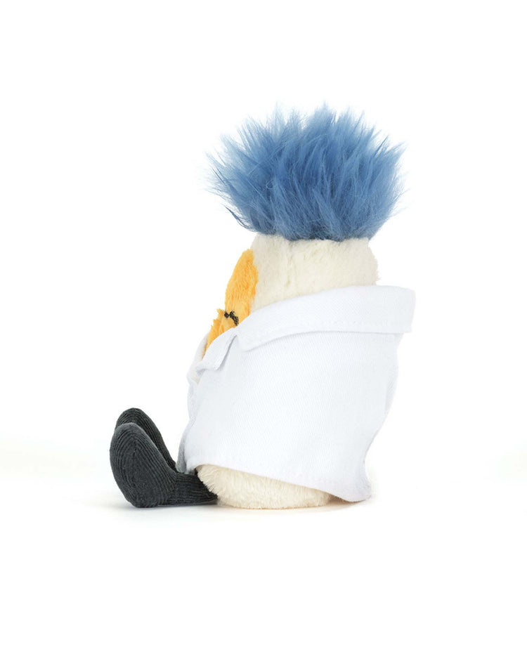 A Jellycat amuseables boiled egg in a scientist outfit with blue spiky hair, a white lab coat, and dark-colored feet sits on a white surface, facing sideways to the left.