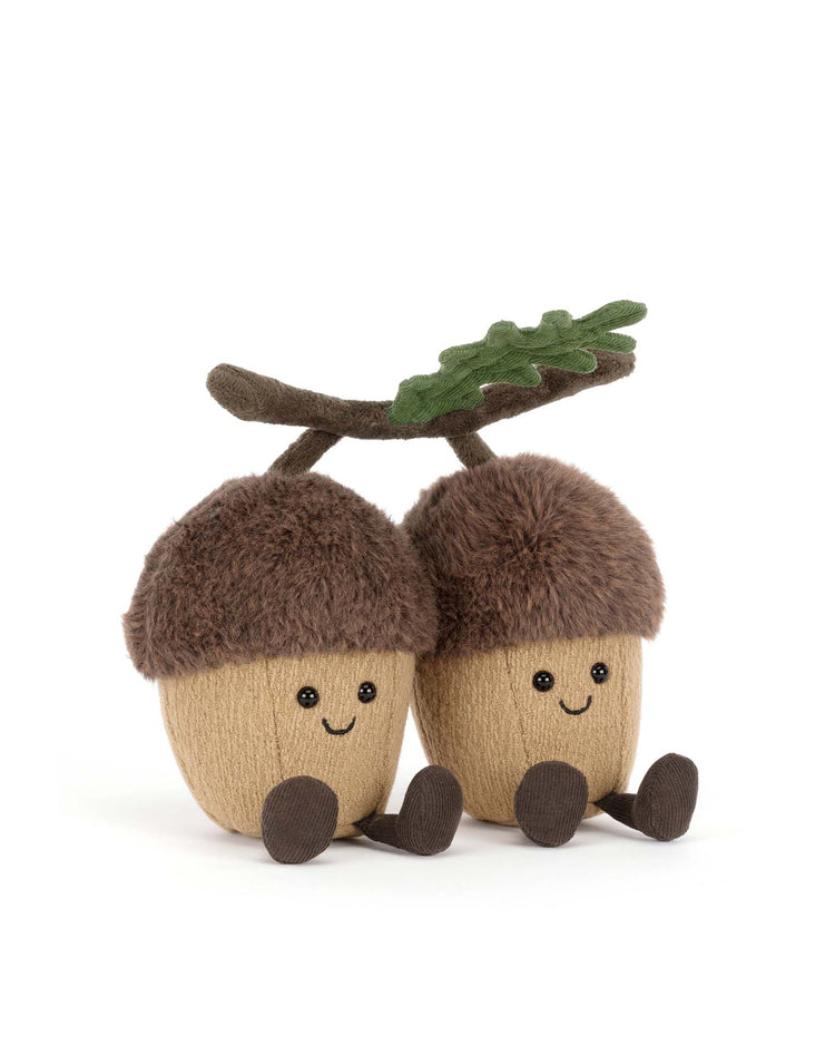 Two Jellycat amuseables Ort & Tum Acorns plush toys with smiling faces, fuzzy brown caps, and little arms and legs sit side by side, connected by a soft branch with a green leaf, against a white background.