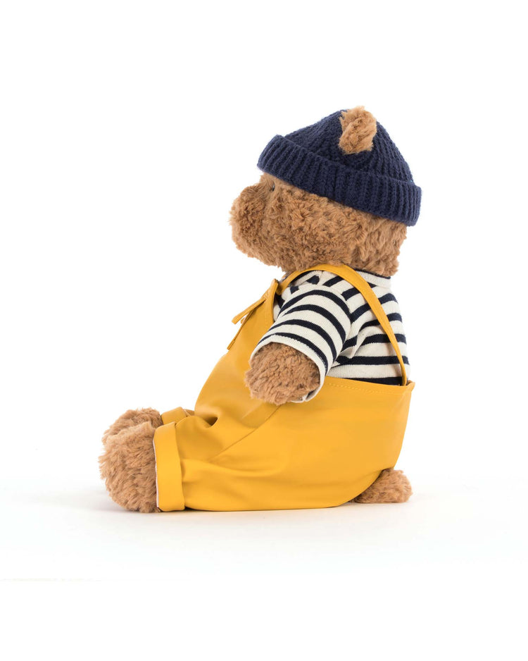 Bartholomew Bear Seafarer Outfit by Jellycat features Bartholomew facing left in a navy knit hat, black-and-white striped shirt, and bright yellow overalls—ready for adventure against a plain white background.