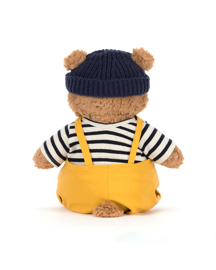 Bartholomew Bear wears the Jellycat Seafarer Outfit—a navy knit beanie, striped sweater, and yellow overalls—and sits ready for adventure against a white background.