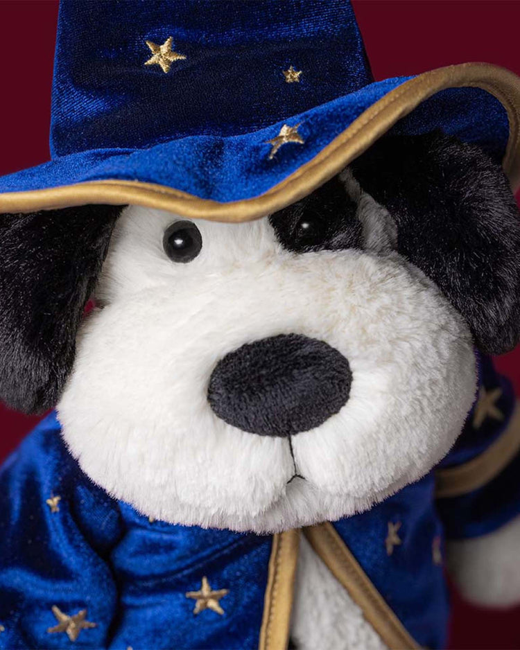 A Jellycat rufferty puppy wizard outfit plush toy, featuring a cuddly dog dressed in a blue hat and robe adorned with gold stars, is displayed against a dark red background.