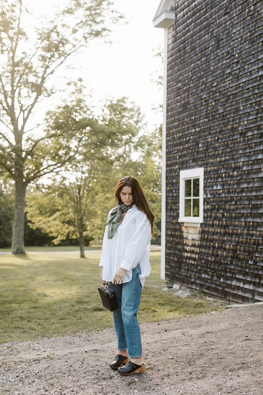 A woman in a white blouse, jeans, and black shoes, carrying a black bag and wearing a green scarf, standing beside a shingled building.