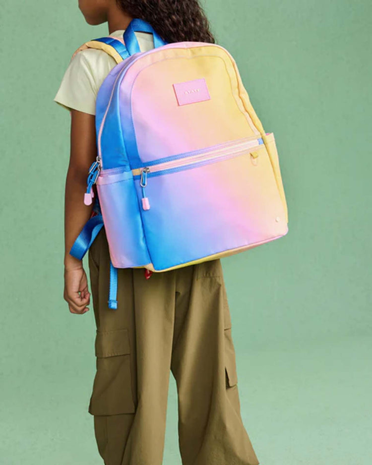 A person wearing a yellow t-shirt and olive pants carries the state bags kane backpack in a blue, pink, and yellow gradient against a green background.
