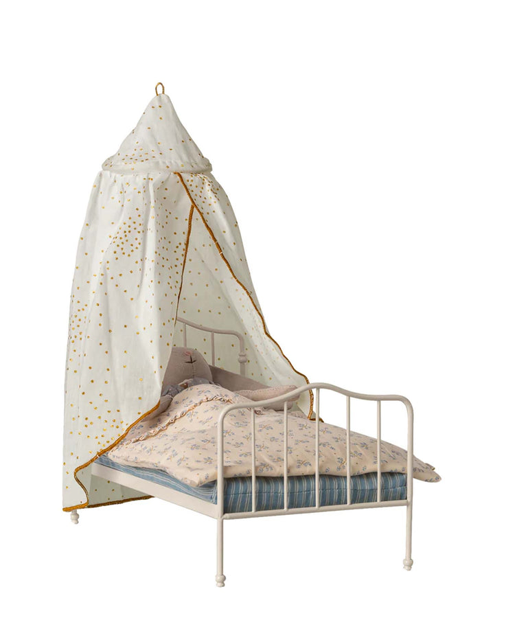 A charming dollhouse scene featuring a small white Maileg metal bed with blue-striped bedding, a floral pillow and comforter, and the Maileg miniature bed canopy in off white draped above.