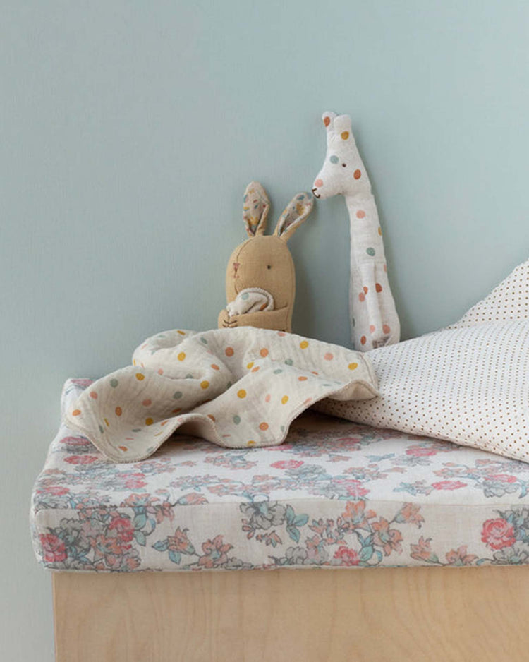 The Maileg lullaby friends, bunny, mini in cream yellow sits on a bed with floral sheets and a polka dot blanket—ideal as a newborn gift or cuddly companion—against a light blue wall.
