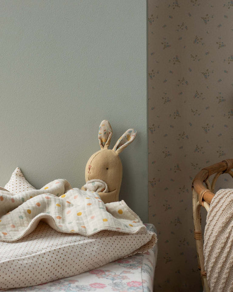 The maileg lullaby friends, bunny, mini - cream yellow plush with long ears and a cuddle cloth peeks out from under a cream polka-dot blanket on a child's bed, beside a chair and delicate floral wallpaper.