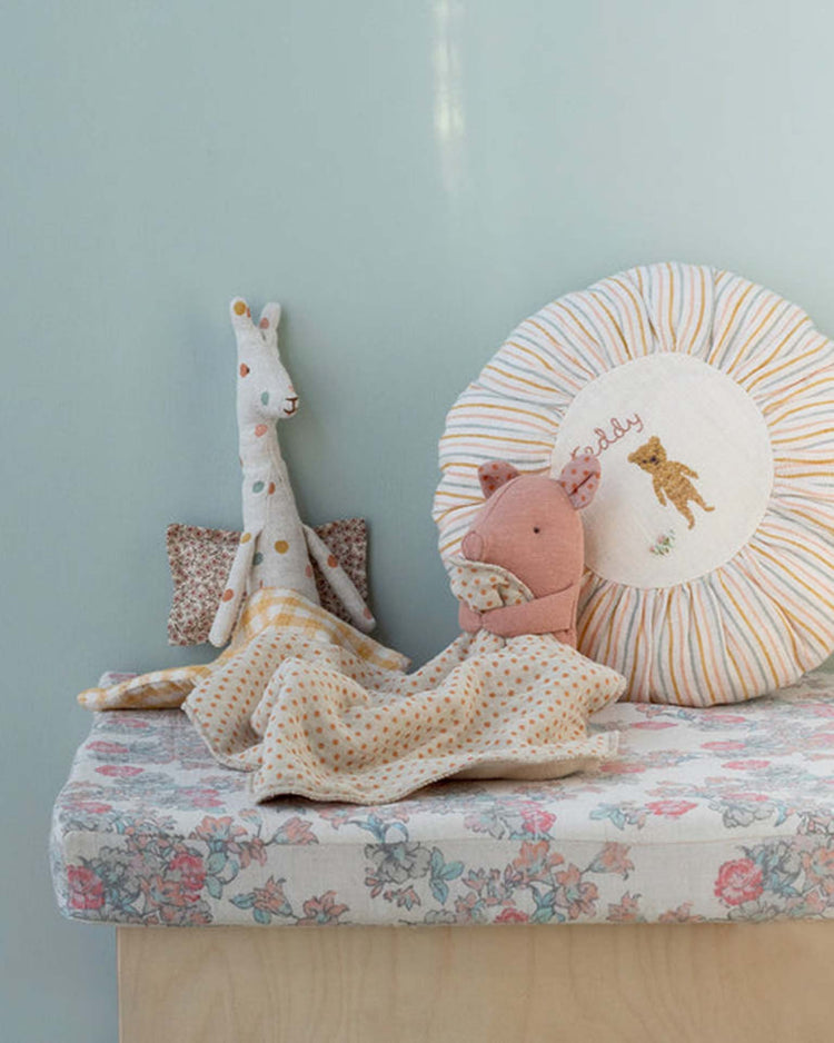Two Lullaby Friends, Pig, Mini - Rose soft toy dolls from Maileg sit on a floral blanket beside a round “baby” pillow with a bear, creating a charming newborn gift against the pale blue wall.
