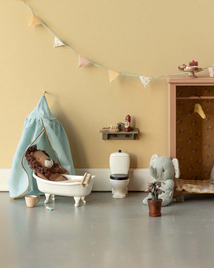 A playful Maileg scene features the little leo lion toy in the bathtub, with a toy elephant on the floor, a mini toilet, cabinet, shelves, pastel bunting above, and a toy plant for a cozy touch.