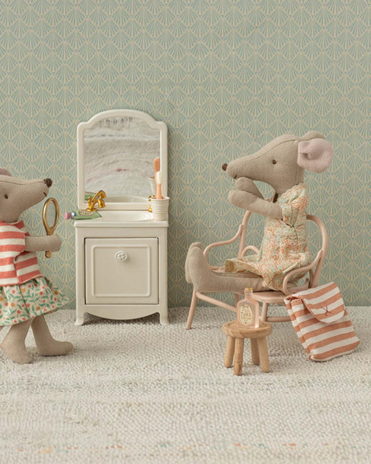 Two mouse figures from the Maileg toothbrush set in bag, coral stripe, enjoy a bedtime routine—one at the vanity with a brush and the other holding a mirror. The pastel colors and patterned wallpaper add charm to this miniature bathroom scene.