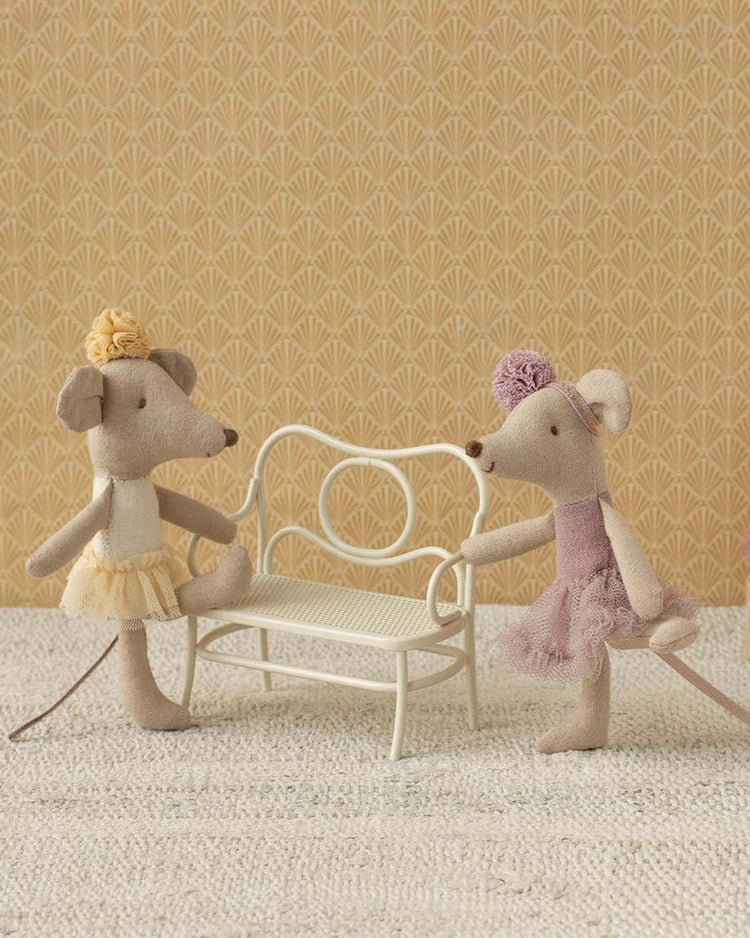 Two ballet toys—a ballerina mouse in a yellow tutu and the maileg little sister ballerina mouse in off white—stand on either side of a small white bench against a patterned beige background.