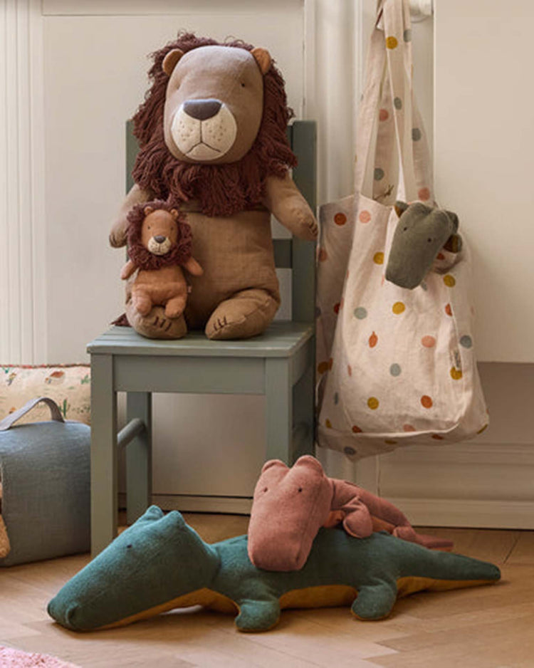 A big crocodile in forest green from Maileg's Safari Friends lies cozy on the wooden floor, with a smaller pink crocodile on its back, while two plush lions rest together on a green chair beside a polka dot tote bag.