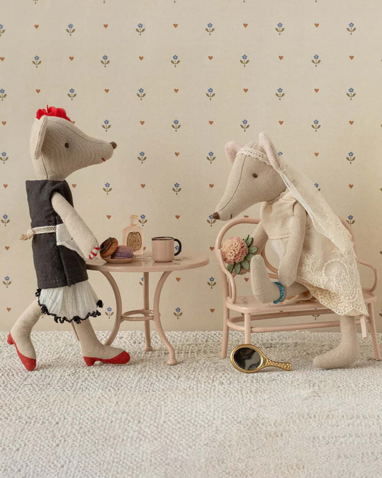 Two Maileg fabric mouse dolls sit at a small table with tea and pastries. One wears the maileg wedding dress for mum mouse, bridal accessories, and holds flowers next to a mirror; the other dons a black dress with red shoes.