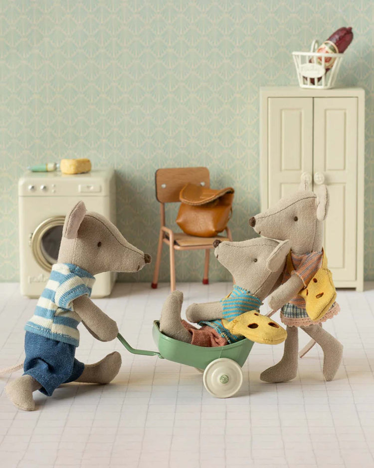 The maileg little sister mouse with bag plays in a miniature room, pushing two others in a green wheelbarrow. The scene includes a washing machine, chair, cabinet, and basket in the background.