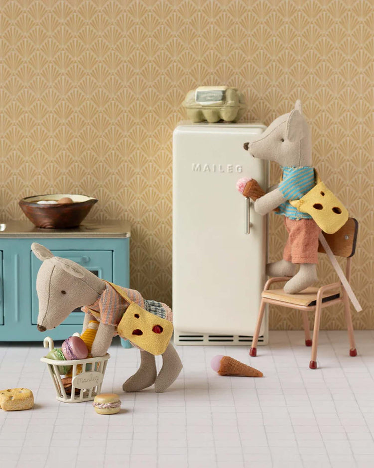 Two little sister mice dressed in Maileg's "clothes and bag for little sister mouse" play in a vintage-style kitchen—one stands on a chair to open the fridge while the other pushes a basket, surrounded by pretend food.
