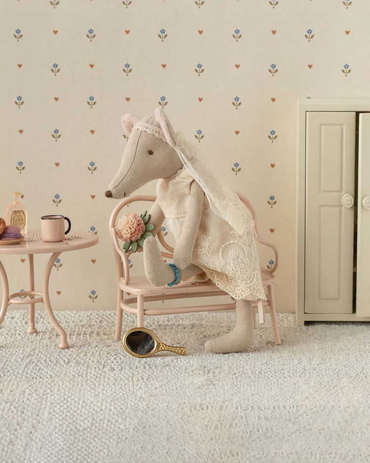 The maileg wedding dress for mum mouse features a charmingly dressed mouse seated on a pink chair with a bouquet. Nearby are a table with cup and bottle, wardrobe, and handheld mirror, all against flower-patterned wallpaper.