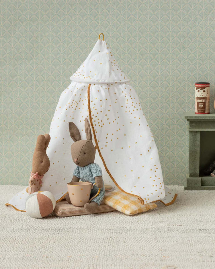 Two plush rabbits, including a Maileg bunny in blue, sit in front of a Maileg miniature bed canopy in off white with gold speckles. Nearby lie a yellow blanket, cup, and ball; behind them is a green shelf holding a small cup and can.