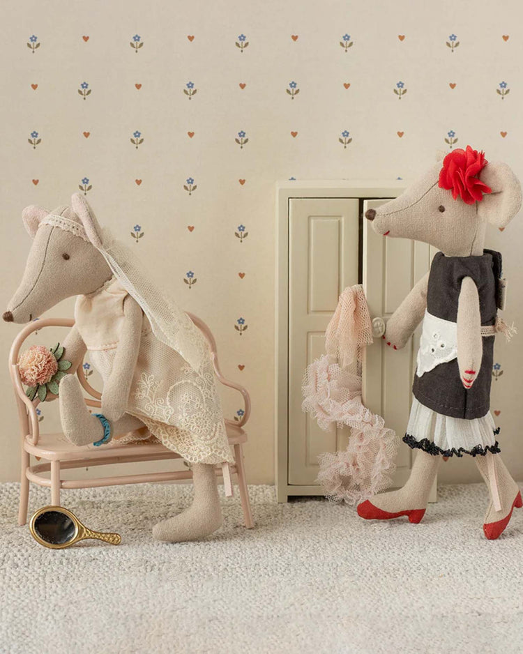 Two stylish Maileg mice are dressed for a wedding. One wears a lace veil, holds flowers, and sits on a pink bench. The other is the Beautician Mum Mouse from Maileg in a black dress, red shoes, and pink scarf, standing nearby.