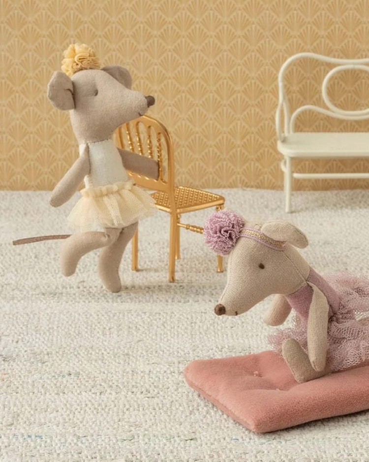 The maileg little sister ballerina mouse in off white poses by a gold chair, while another plush ballerina mouse sits on a pink mat near a white bench, set against patterned wallpaper.