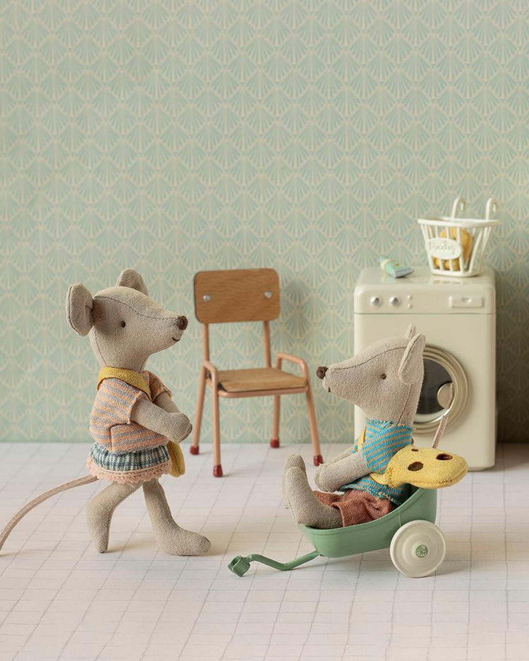 A maileg little sister mouse with bag and plush mouse friend play with a green wheelbarrow in a room featuring a wooden chair, toy washing machine, and basket against patterned wallpaper.