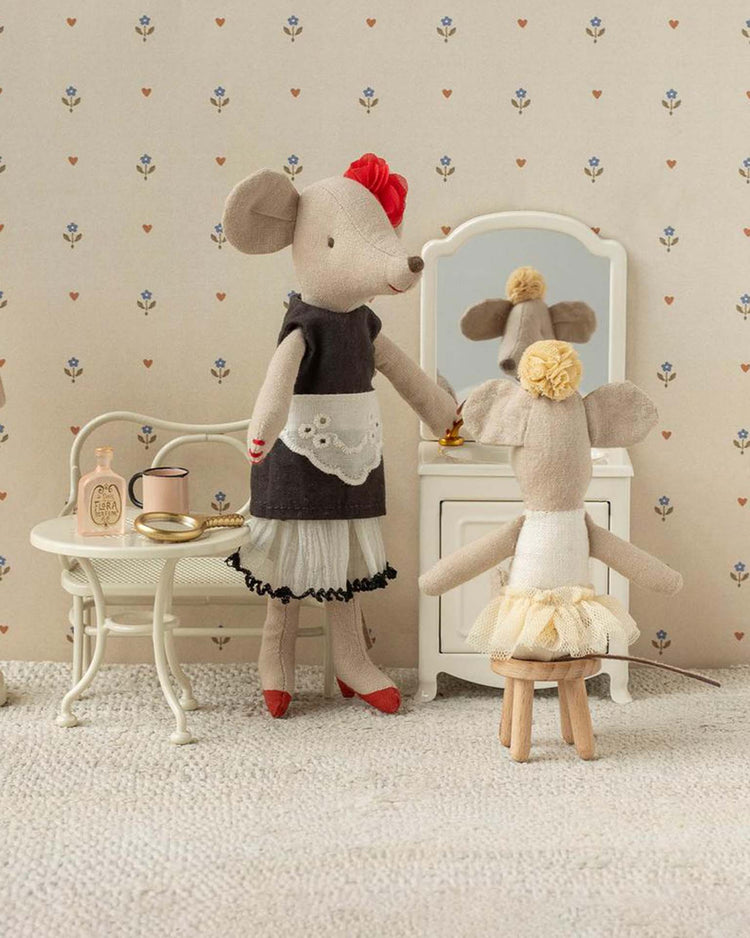 A Maileg little sister ballerina mouse in off white sits on a stool at the dollhouse vanity, gazing in the mirror, while her older sister in a black dress stands nearby. Accessories rest on a small table close to the mice.