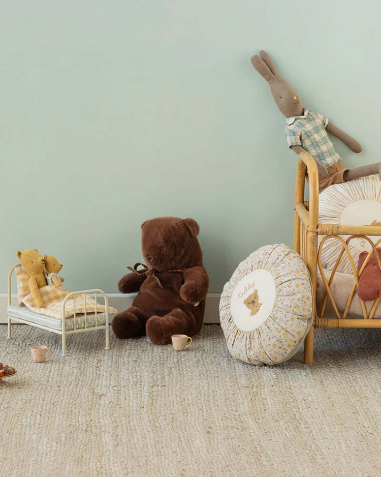 A cozy nursery showcases a maileg medium teddy bear plush in dark chocolate, nestled among toy teacups, a round floral pillow, two cuddly stuffed bears, and a plaid rabbit resting in a rattan crib against a pastel blue wall.
