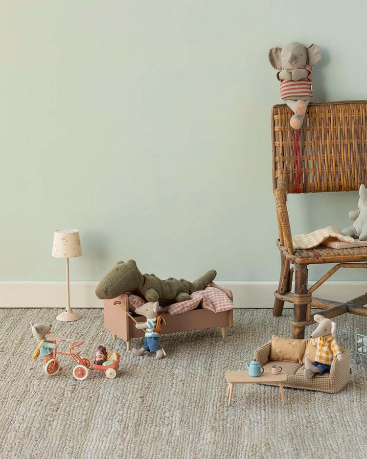 A cozy playroom with plush animal toys: a crocodile in bed, mice on a tricycle and sofa, and Maileg’s Little Jumbo elephant with magnetic hands on a wicker chair. A glowing lamp highlights the soft, neutral-toned setting.