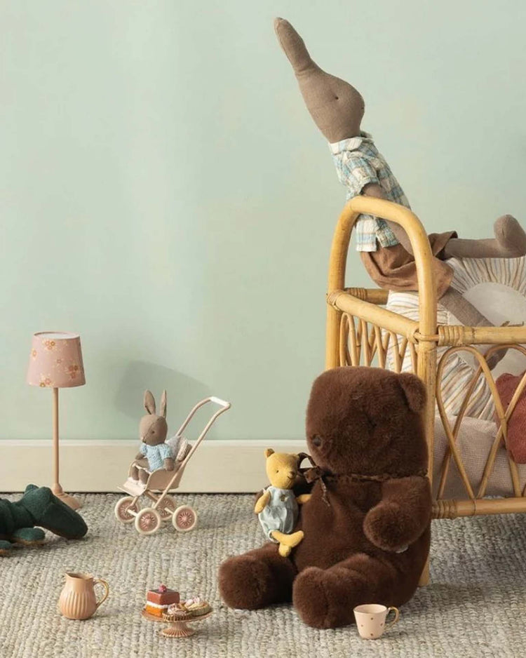 A cozy nursery scene with a Maileg medium teddy bear plush in dark chocolate, holding a small yellow bear, plus a soft plush rabbit in a crib, another rabbit in a toy stroller, mini tea set, and lamp on a textured carpet.
