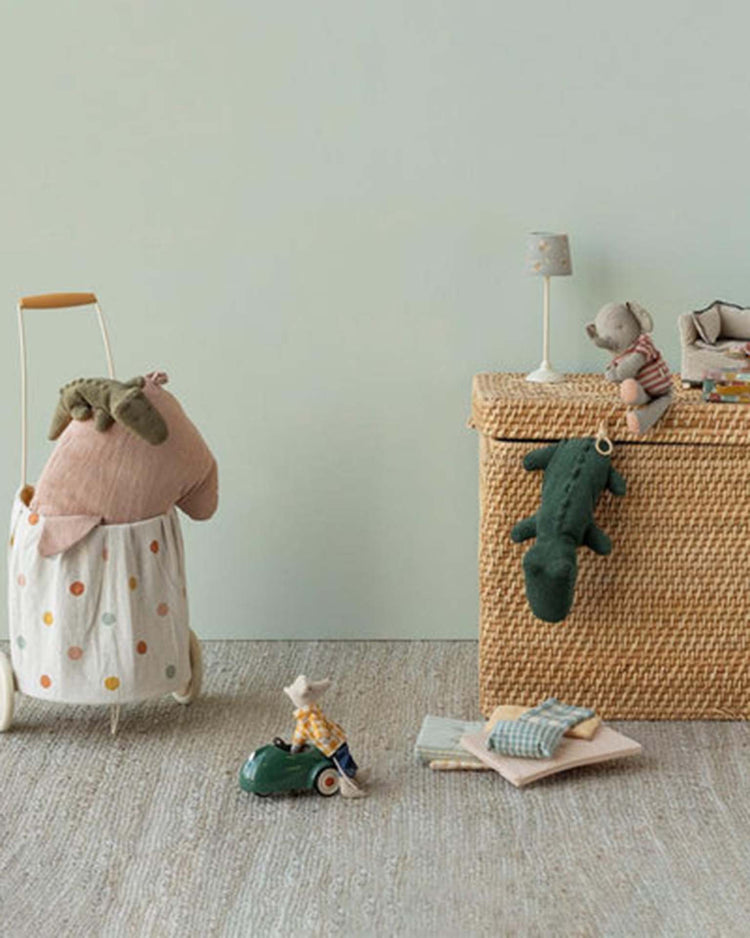 A nursery scene features Maileg's small crocodile in forest green from Safari Friends on a wicker basket, with soft toys and playful animals arranged atop a neutral, textured floor.