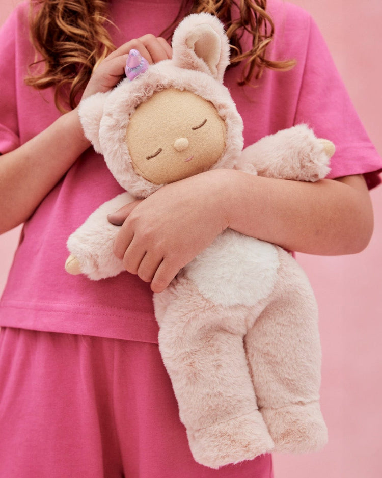 A child in pink pajamas hugs the cozy dinkum unicorn sugarpie pink by dinkum dolls—this huggable stuffed animal with closed eyes wears a fuzzy unicorn costume and stands against a pink background.