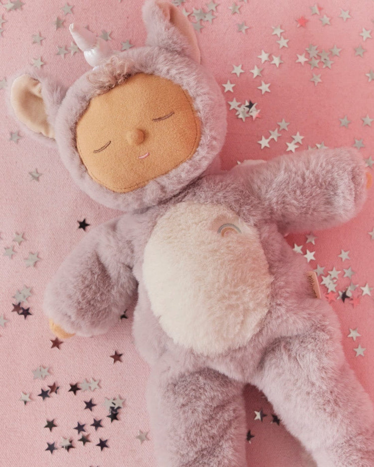 The cozy dinkum unicorn sweetpea lilac by dinkum dolls features a soft unicorn companion in a lilac costume with a horn, resting on a pink surface and surrounded by silver star confetti.