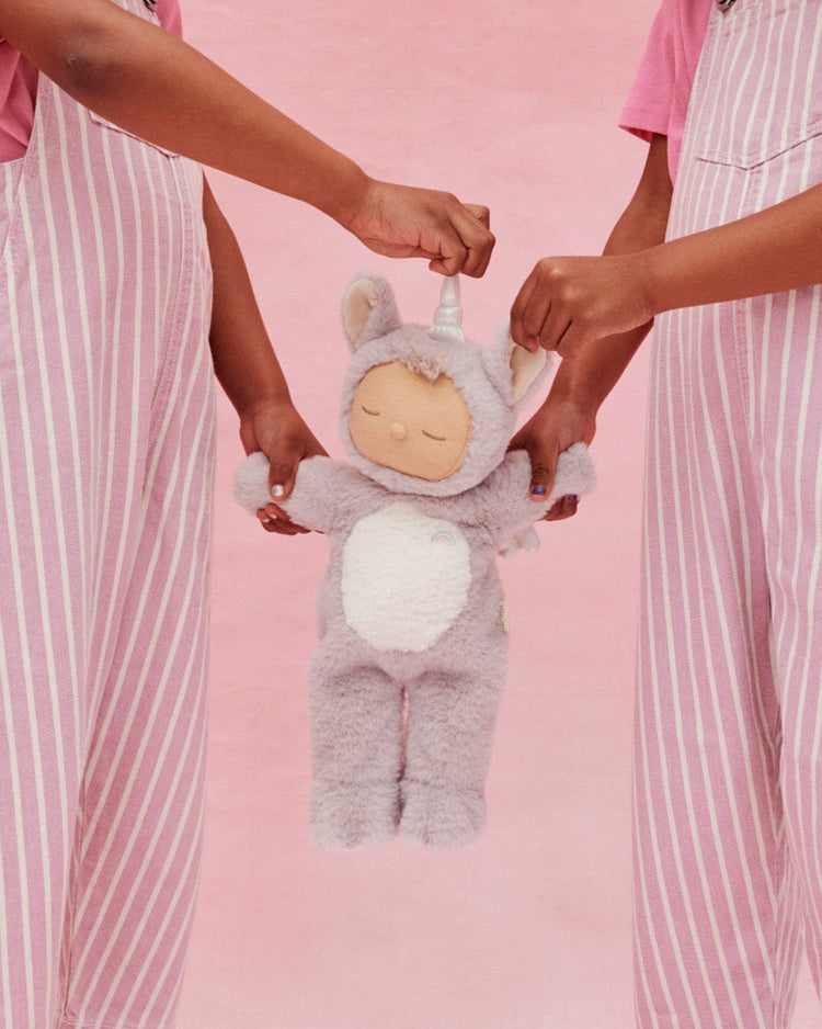 Two people in pink and white striped overalls hold the dinkum dolls cozy dinkum unicorn sweetpea lilac, a plush doll with a soft grey body, white belly, closed eyes, and a small horn, set against a pink background.