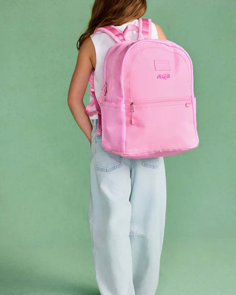 A child wearing a white sleeveless top and light blue jeans stands facing away, carrying the Kane Double Pocket Backpack in Pink by State Bags—a perfect kids’ school backpack—against a green background.