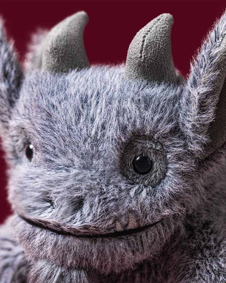 A close-up of the Jellycat Growlston Gargoyle plush—a fuzzy grey creature featuring two soft horns, pointed ears, black button eyes, and a gentle smile—set against a deep red background.