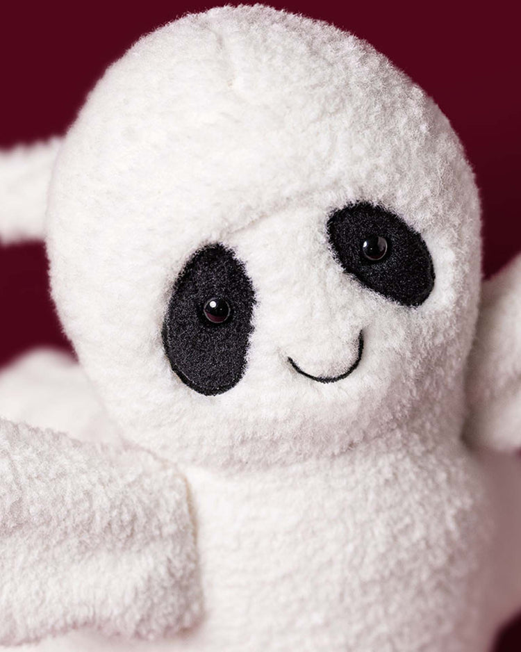 A close-up of a Jellycat Ghost Bob plush toy with soft white fur, black oval patches around its button eyes, and a stitched smiling mouth, set against a burgundy background.