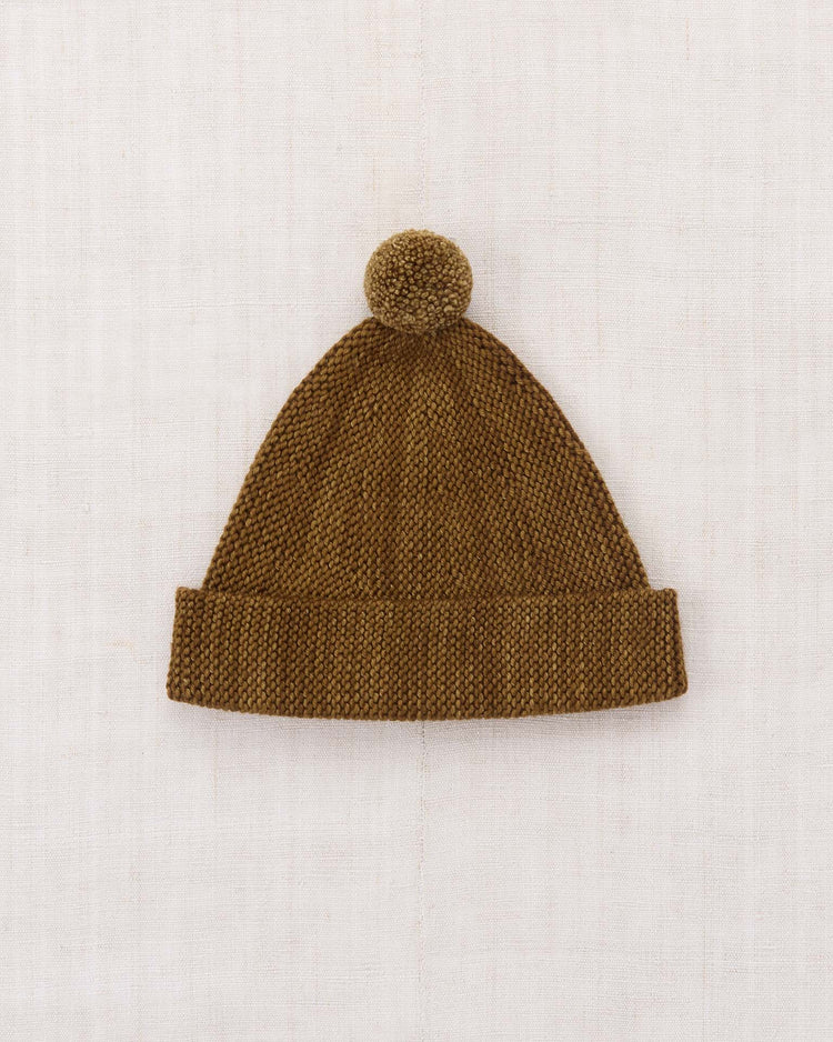The garter hat in nutmeg by misha + puff, crafted from brown Merino wool with a folded, adjustable brim and a pom-pom on top, is laid flat against a light, textured background.