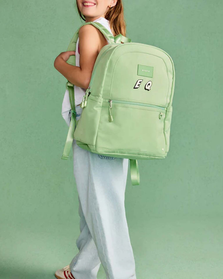 A smiling person wearing a white top and light jeans stands sideways holding a large light green STATE Bags Kane Double Pocket Backpack decorated with the letters E Q, set against a green background.
