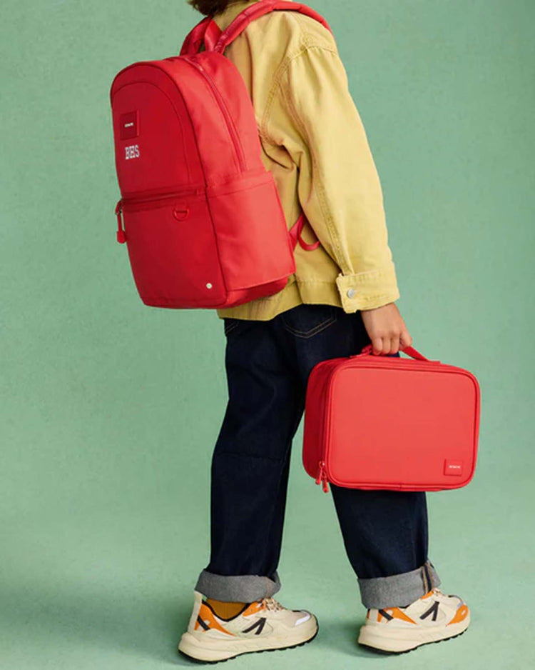 A child wearing a yellow jacket, dark jeans, and sneakers stands against a green background, carrying the STATE Bags Kane Backpack in red and holding a matching red lunchbox.