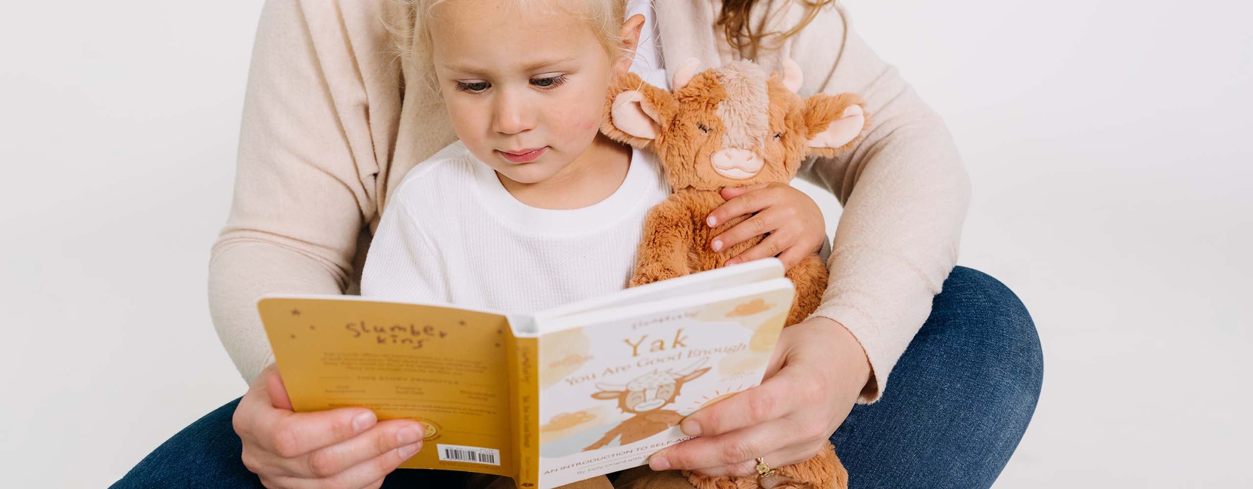 A child holding a stuffed animal attentively reads a book with an adult's assistance.